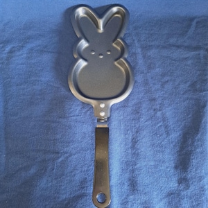 Könnte beinhalten: Eine silberfarbene, hasenförmige Pfannkuchenform mit einem langen Griff aus Edelstahl. Die Form zeigt ein detailliertes Hasengesicht mit zwei Ohren und drei kleinen Punkten für Augen und Nase. Der Griff hat eine Schlaufe zum Aufhängen.