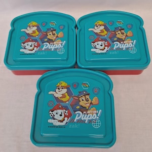 Op de afbeelding: Drie turquoise en rode broodtrommels in sandwichvorm. Elke broodtrommel heeft de tekst "PLAYTIME Pups! zak!" en illustraties van cartoonpuppy's. De deksels zijn turquoise en de bodems rood.