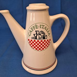 Puede incluir: Cafetera de cerámica blanca con boquilla larga y asa. La cafetera presenta el texto "CAFFÈ ITALIA" en verde, junto con un diseño a cuadros rojos y blancos e ilustraciones de comida y bebida.