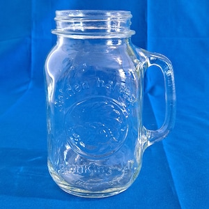 Puede incluir: Tarro de vidrio transparente para beber con asa. El tarro tiene las palabras "golden harvest" y "drinking jar" en relieve en el lateral. El tarro está vacío y se encuentra sobre una superficie azul.