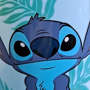 May include: Close-up of a blue and teal image featuring the character Stitch from Lilo & Stitch. The character's face is centered, with large blue eyes, a dark blue nose, and a small smile. The background has a light blue color with teal leaf patterns.