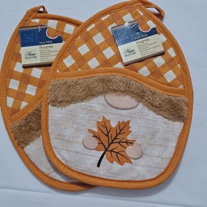 May include: Two orange and white checkered pot mitts with a gnome design. The gnome has a faux fur beard and a leaf design. Each mitt has a tag that reads "Pot Mitt" and "Home Collection".