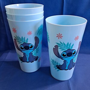 May include: A set of four light blue plastic tumblers featuring a cartoon character design. Each tumbler has a printed image of a blue alien with large ears, surrounded by pink flowers and green leaves.