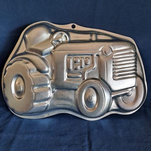 May include: A silver metal tractor-shaped cake pan. The pan has a detailed design with a front grill, wheels, and a steering wheel.