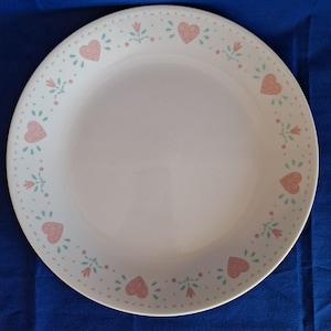 Puede incluir: Plato de cerámica blanco con un borde de corazones rosas y pequeños diseños florales en rosa y azul claro. El plato tiene un diseño sencillo y limpio, adecuado para el uso diario.
