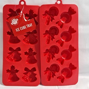 Könnte beinhalten: Zwei rote Silikon-Eiswürfelformen. Eine Form hat Glocken- und Stechpalmen-Designs, die andere hat Zuckerstangen- und Bonbonformen. Ein Etikett lautet "ICE CUBE TRAY". Ideal für Getränke mit Weihnachtsmotiven.