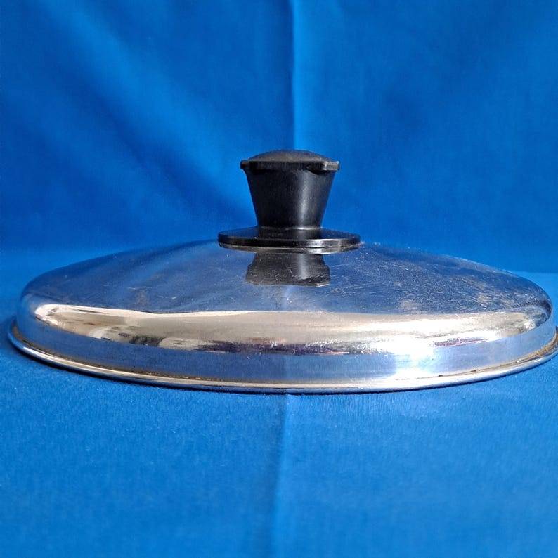 May include: A stainless steel pot lid with a black handle. The lid is round with a shiny, reflective surface. The handle is cylindrical and sits atop the lid's center. The background is a solid blue.