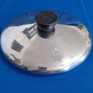 May include: A shiny, silver-colored metal lid with a black, round handle. The lid reflects the surrounding environment, creating a mirrored effect. The handle is made of a dark, possibly rubbery material.