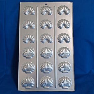 May include: A silver metal baking mould featuring a seashell design. The mould has 18 individual shell-shaped cavities, arranged in three columns and six rows. A small hole is at the top for hanging.