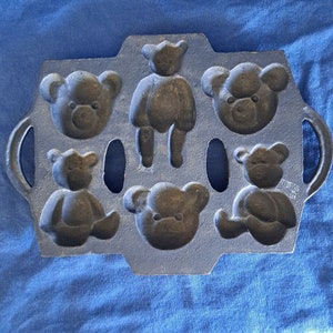 May include: A cast iron baking pan with six bear-shaped moulds. The pan is dark grey and has two handles on the sides.