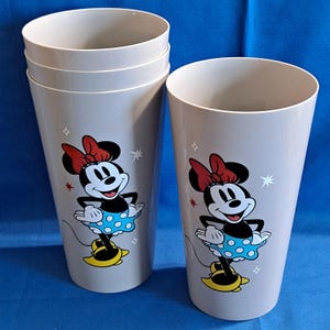 May include: Six grey plastic tumblers with a Minnie Mouse design. Each tumbler features a cartoon image of Minnie Mouse wearing a red bow, blue polka-dot skirt, and yellow shoes. The tumblers are decorated with star accents.