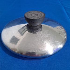 Puede incluir: Una tapa de olla plateada y brillante con un asa negra y redondeada. La tapa tiene forma circular y un acabado de metal cepillado. El asa es de un material oscuro, posiblemente de goma. La tapa está sobre una superficie azul.