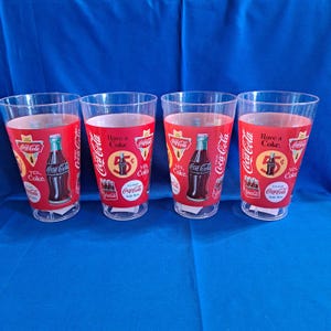 May include: Four clear plastic Coca-Cola glasses. Each glass has a red band with Coca-Cola branding, a bottle illustration, and the text "Have a Coke." The glasses are on a blue surface.