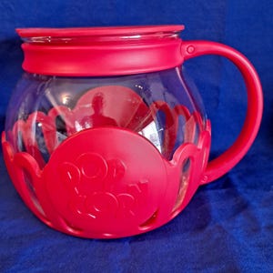 Op de afbeelding: Rode glazen popcornmaker met een doorzichtig deksel en een handvat. De maker heeft een rode basis met de woorden "Pop Corn" erop gegraveerd.