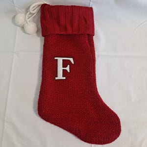 Può includere: Una calza di Natale rossa a maglia con la lettera "F" bianca sul davanti. La calza ha un polsino a coste e un cordino bianco con due piccoli pompon bianchi. Per decorazioni natalizie.
