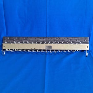 May include: A rectangular tie rack with a dark grey textured top and a silver-coloured metal base. The rack has multiple hooks for hanging ties and is set against a blue background.
