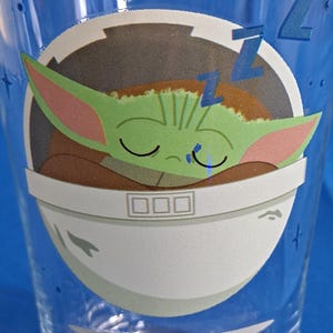Peut inclure: Un verre &agrave; boire bleu transparent avec un dessin d'un personnage extraterrestre vert endormi avec de grandes oreilles, reposant dans une nacelle blanche. Le motif comprend des Z bleus et un fond blanc, avec un motif d'&eacute;toiles bleues sur le verre.