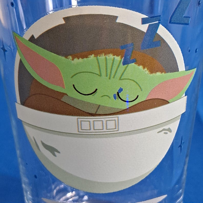 Peut inclure: Un verre &agrave; boire bleu transparent avec un dessin d'un personnage extraterrestre vert endormi avec de grandes oreilles, reposant dans une nacelle blanche. Le motif comprend des Z bleus et un fond blanc, avec un motif d'&eacute;toiles bleues sur le verre.