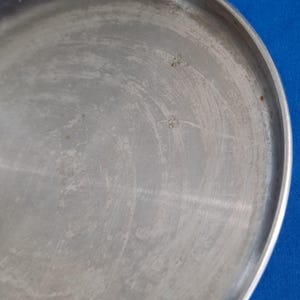 May include: A close-up of a round, silver-colored metal tray with a slightly raised edge. The surface shows visible scratches and imperfections, with a few small, dark spots. The tray is set against a blue background.