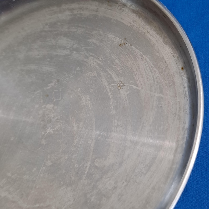 May include: A close-up of a round, silver-colored metal tray with a slightly raised edge. The surface shows visible scratches and imperfections, with a few small, dark spots. The tray is set against a blue background.