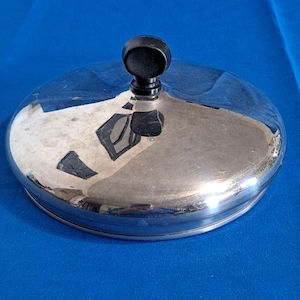 May include: A shiny, silver-colored lid with a black knob. The lid is round and reflective, with a slightly domed shape. The knob is small and circular, and is attached to the center of the lid. The lid is sitting on a blue surface.