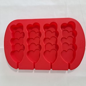 May include: A red silicone mold for making heart-shaped lollipops or candies. The mold has four rows of heart shapes, each with a slot for a stick. The brand name "Wilton" is printed on the side.