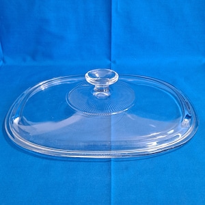 May include: A clear glass oval-shaped lid with a small, round handle. The lid has a raised, circular design in the centre with vertical lines radiating outwards. The edges are smooth and rounded, suitable for cookware.