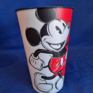 Puede incluir: Un vaso de metal blanco y rojo con un dise&ntilde;o de Mickey Mouse sonriente. El vaso tiene un borde negro y una ilustraci&oacute;n de Mickey Mouse en blanco y negro. El fondo es azul liso.
