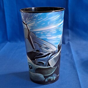May include: Tall plastic tumbler featuring a blue and white space scene with a spaceship and a character from a popular science fiction series. The tumbler has a black rim and a detailed graphic design.