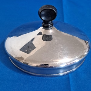 May include: A circular, polished silver-tone metal lid featuring a smooth, domed surface and a central, rounded black knob handle. The metallic finish is reflective, showing light highlights across the curved top and a defined rim edge.