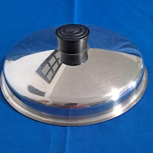 May include: A shiny, stainless steel lid with a black, rounded handle. The lid reflects the surrounding environment, showing a window. The lid is circular and appears to be for a pot or pan.