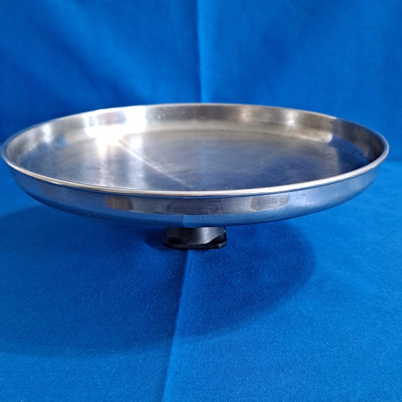 May include: A stainless steel cake stand with a round, shallow tray and a small, black base. The stand is set against a blue background, and the metal surface reflects light.