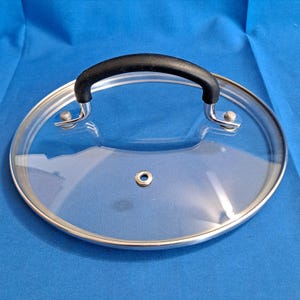 May include: A clear glass pot lid with a stainless steel rim and a black handle. The lid has a small hole in the center and is designed to fit a pot. The handle is curved for easy gripping.