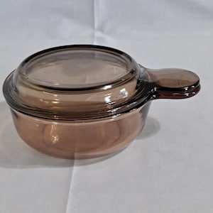 May include: A brown glass casserole dish with a matching lid. The dish has a single handle and is made of transparent glass. The lid fits snugly on top of the dish. The dish is sitting on a white surface.