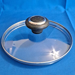 May include: A circular tempered glass lid with a silver-toned metal rim and a black heat-resistant knob. The center of the knob features the embossed text The Pampered Chef. The lid is transparent, allowing for a clear view through the glass surface.