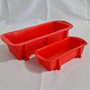 May include: Two red silicone loaf pans of different sizes. The larger pan has a rectangular shape with rounded edges and a flat base. The smaller pan has a similar design. Both pans are made of a flexible material and are suitable for baking.