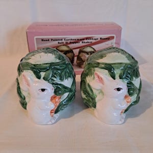 May include: A set of two hand-painted ceramic salt and pepper shakers shaped like cabbages with a white bunny rabbit peeking out of each. The bunnies are holding carrots and the cabbages are green with white accents.