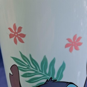 May include: A white mug with a cartoon character design. The mug features pink flower and green leaf designs. The bottom of the mug has a blue cartoon character design.