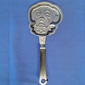 May include: A silver-colored metal pancake mold in the shape of a dog's face. The mold has a long handle with a black grip and a loop at the end for hanging. The dog's features are clearly defined in the mold.