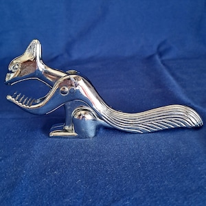May include: A chrome-plated squirrel-shaped nutcracker. The nutcracker features a detailed design with a squirrel's head, body, and tail. The jaws of the squirrel are open, ready to crack nuts. The nutcracker is on a blue surface.