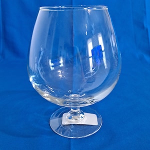 May include: A clear, large brandy snifter glass with a rounded bowl and a short stem. The glass is made of transparent material, reflecting light and the blue background. The base is a flat, circular shape. A white label is attached to the base.