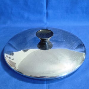 May include: A circular, polished stainless steel pot lid featuring a smooth, reflective surface and a centered, black, conical-shaped handle knob. The lid has a slightly domed profile and a clean, metallic finish throughout its entire top surface.