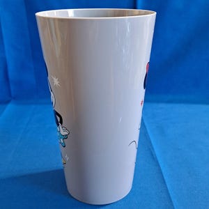 May include: A tall, light grey plastic tumbler featuring cartoon images of Mickey and Minnie Mouse. Mickey is wearing blue shorts and gloves, while Minnie sports a red bow. The tumbler has a wide opening and a slightly tapered shape.