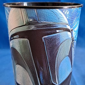 May include: A tall, black-rimmed plastic cup featuring a detailed illustration of a helmet in shades of blue, gray, and black. The design is reminiscent of a science fiction character. The cup has a glossy finish.