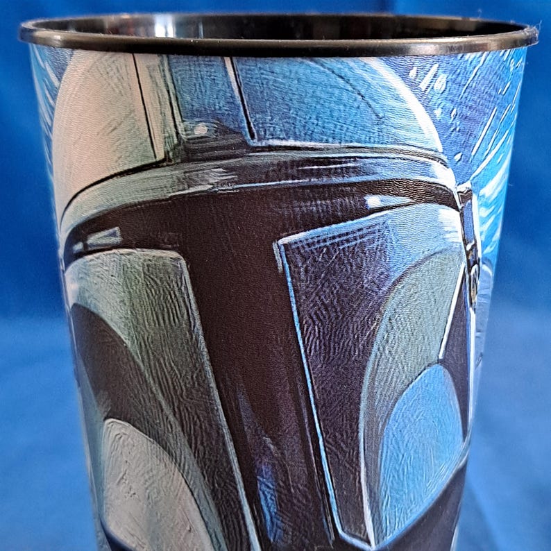 May include: A tall, black-rimmed plastic cup featuring a detailed illustration of a helmet in shades of blue, gray, and black. The design is reminiscent of a science fiction character. The cup has a glossy finish.