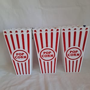 May include: Three white popcorn containers with red vertical stripes and a scalloped top edge. Each container has a red circle with the words "POP CORN" in white. The containers are empty and ready to be filled.