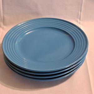 Puede incluir: Una pila de cuatro platos de cerámica azul claro. Cada plato tiene un borde ligeramente elevado y acanalado. Los platos están apilados uno encima del otro, siendo el plato superior totalmente visible. Los platos son de color liso y mate.