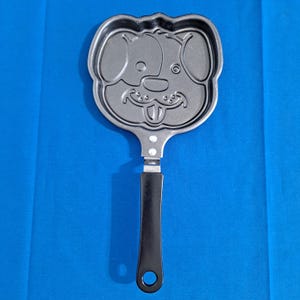 Könnte beinhalten: Eine graue, hundeförmige Pfanne mit schwarzem Griff. Die Pfanne hat ein detailliertes Hundegesicht-Design, einschließlich Augen, Ohren und einem lächelnden Mund. Der Griff hat ein Loch zum Aufhängen. Die Pfanne ist aus Metall.