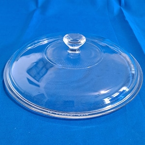 Puede incluir: Una tapa de vidrio transparente con forma redonda y un pequeño pomo transparente en la parte superior. La tapa tiene una serie de anillos concéntricos que irradian desde el centro. El borde de la tapa tiene un borde ligeramente elevado. El fondo es azul liso.