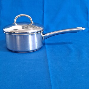 Puede incluir: Una cacerola de acero inoxidable con tapa y mango largo. La olla pequeña tiene un asa curva en la tapa y un asa larga y recta que se extiende desde el lateral. La olla está sobre una superficie azul.